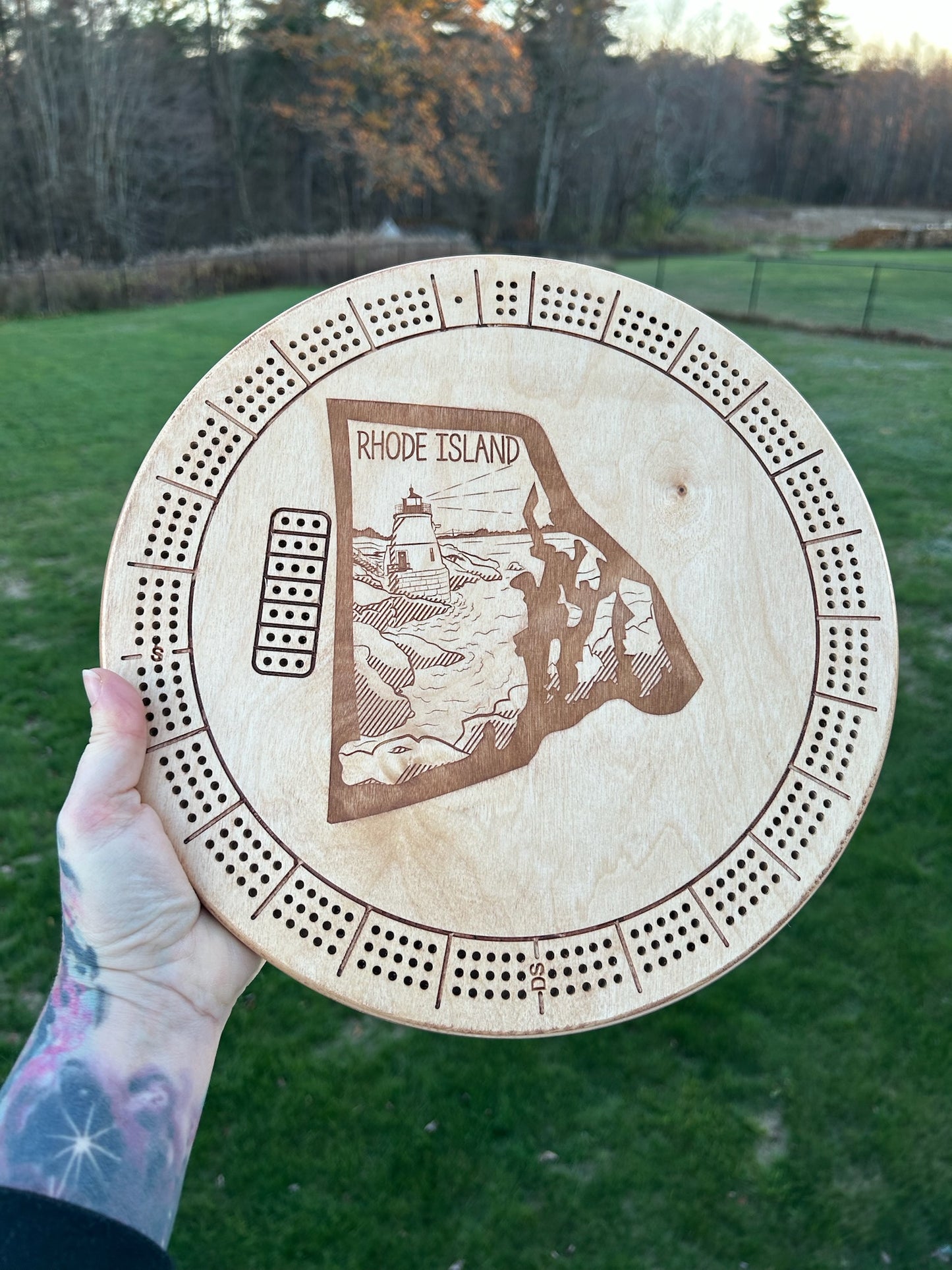 A round cribbage board with the state of Rhode Island motif carving, crafted from solid wood, held against a nature backdrop.