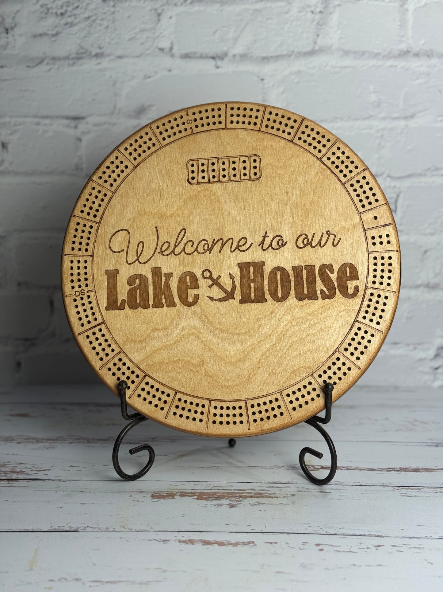 12" round light brown cribbage with 'Welcome to our Lake House' text on a stand against a white brick wall.