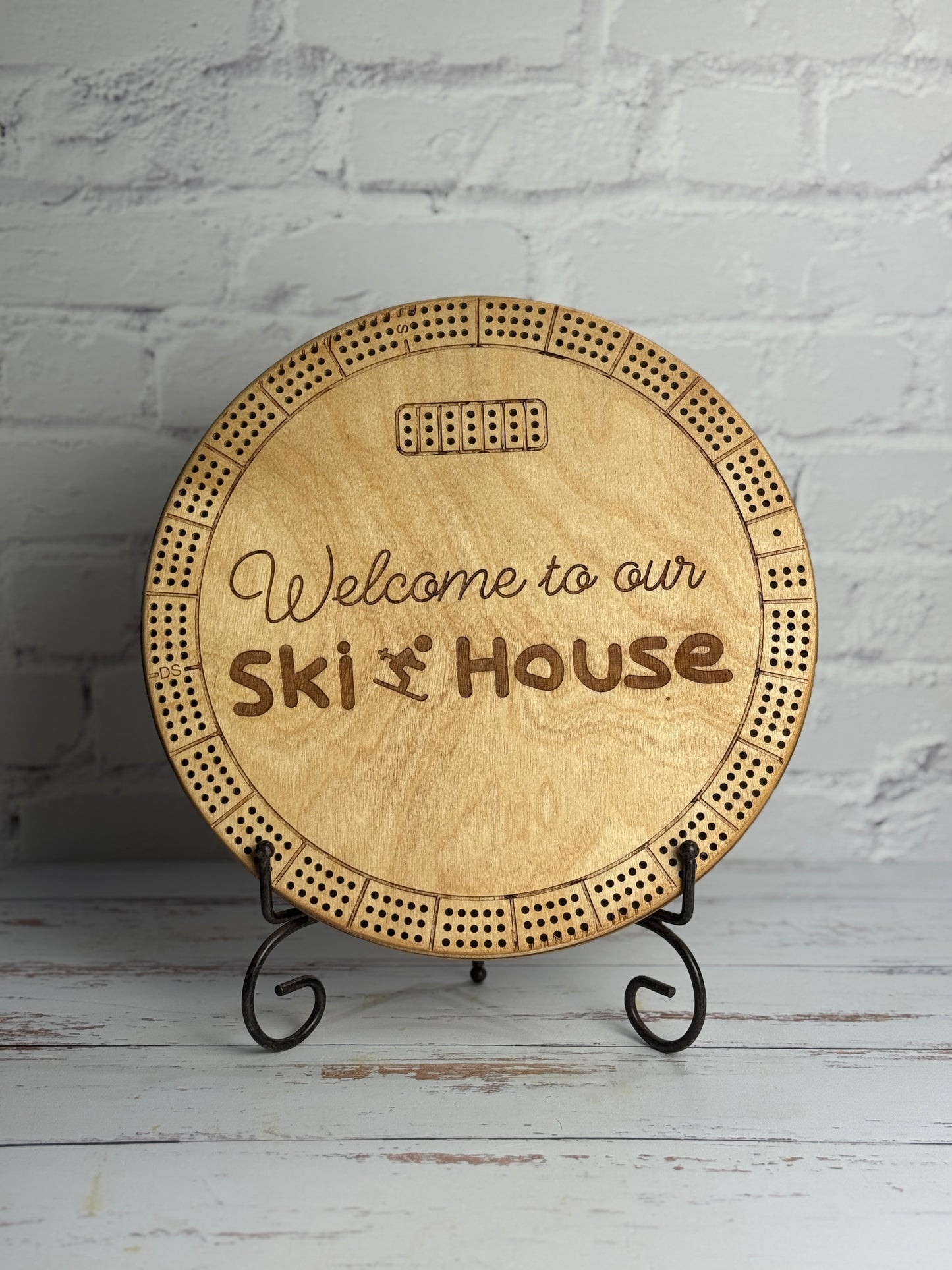 12" round light brown cribbage with 'Welcome to our Ski House' text on a stand against a white brick wall.