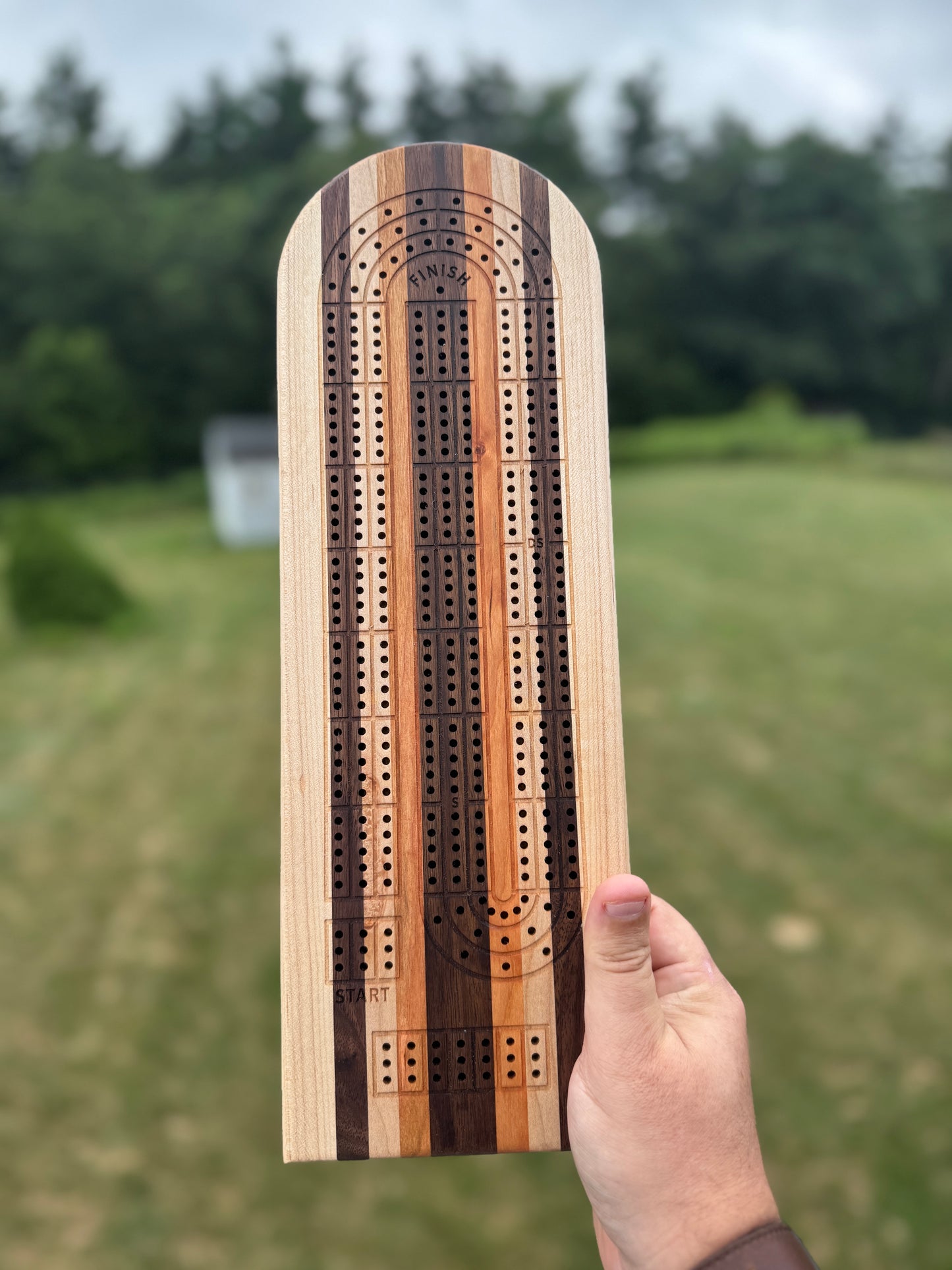 Wooden cribbage board held by a hand with a blurred green outdoor background. Woods are maple, walnut, maple, cherry, walnut, cherry, maple, walnut, maple (left to right).