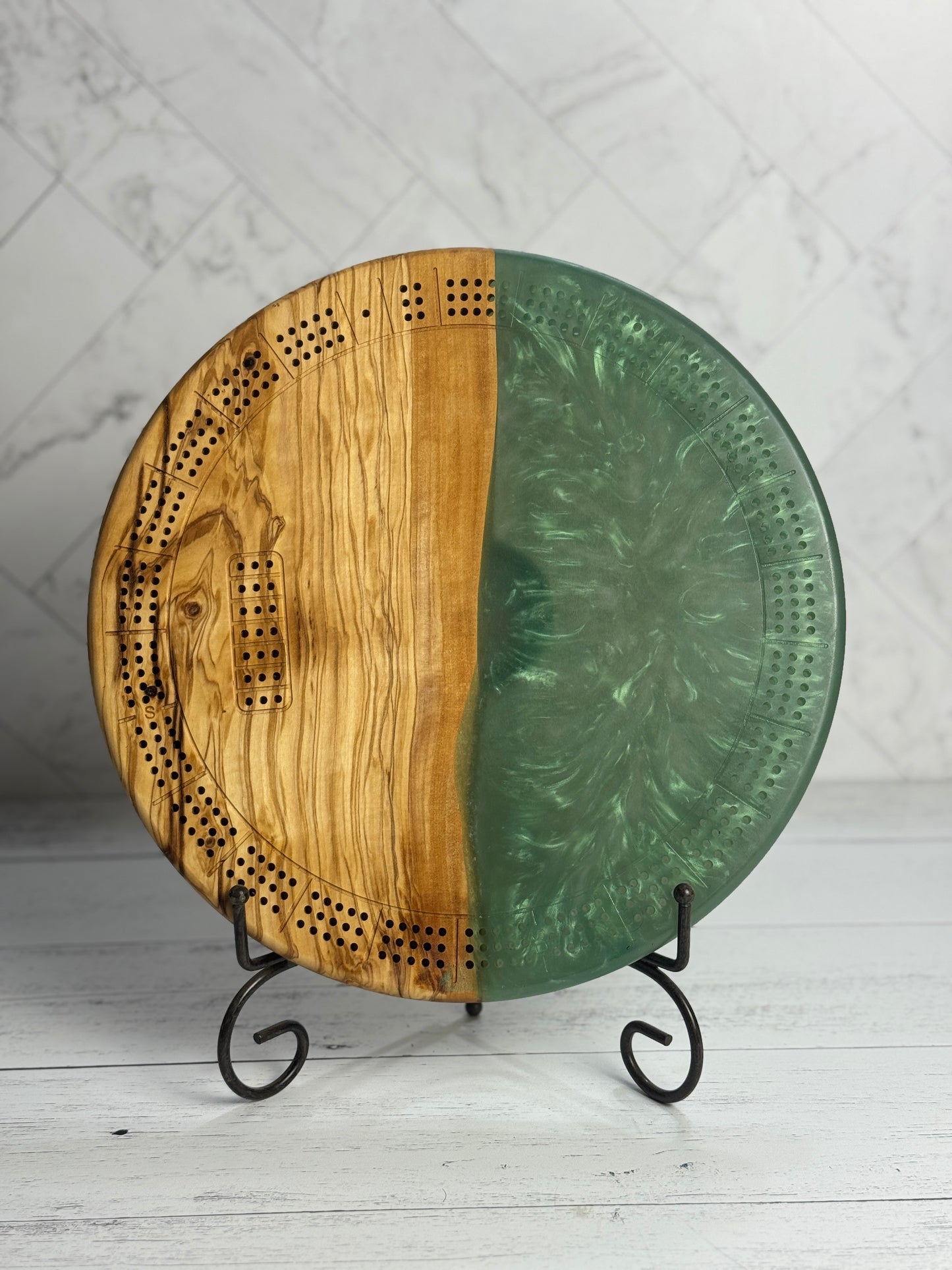 A small round cribbage board with a mix of olivewood and green resin on a stand against a light background.