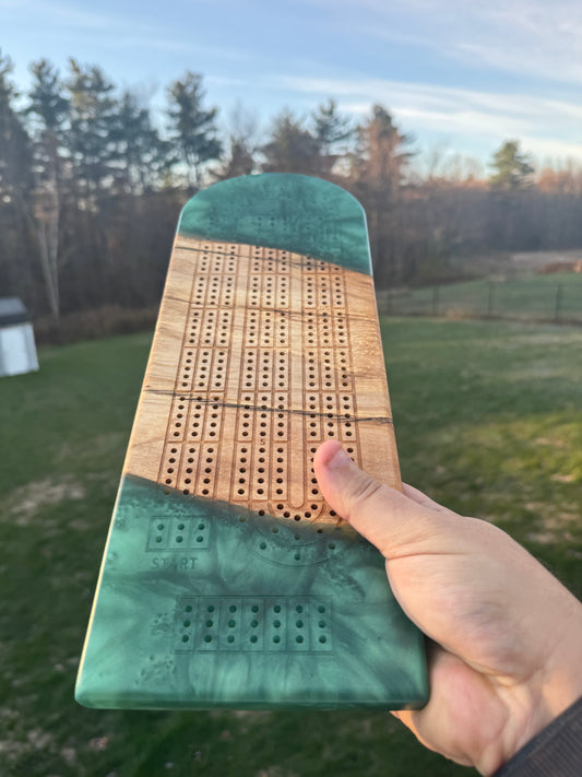Hand holding a green resin rectangular cribbage board, in a paperclip layout, against a nature background