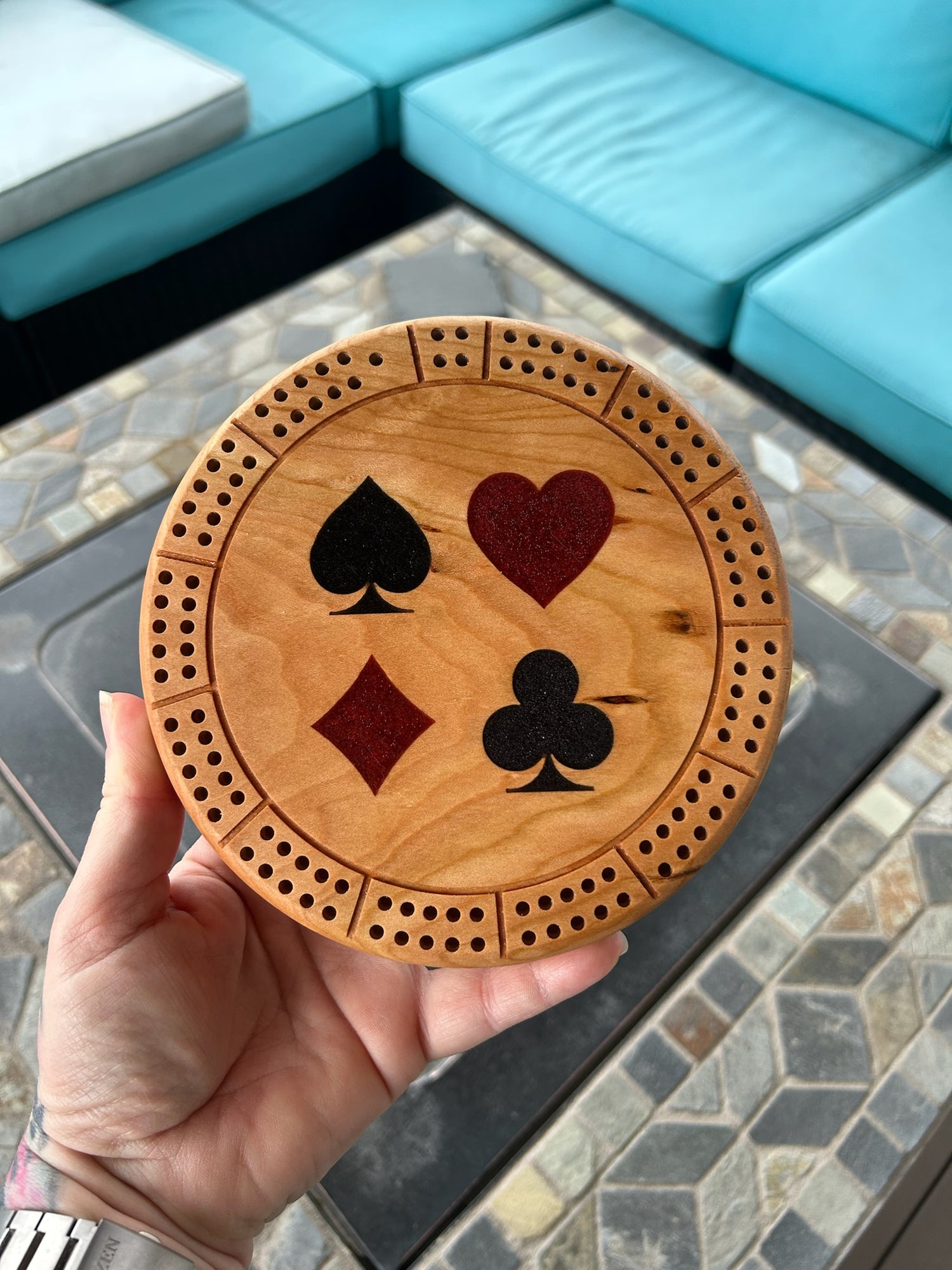 Handcrafted 6" hardwood round cribbage board with resin-filled card suit symbols held by a hand against a stone surface.