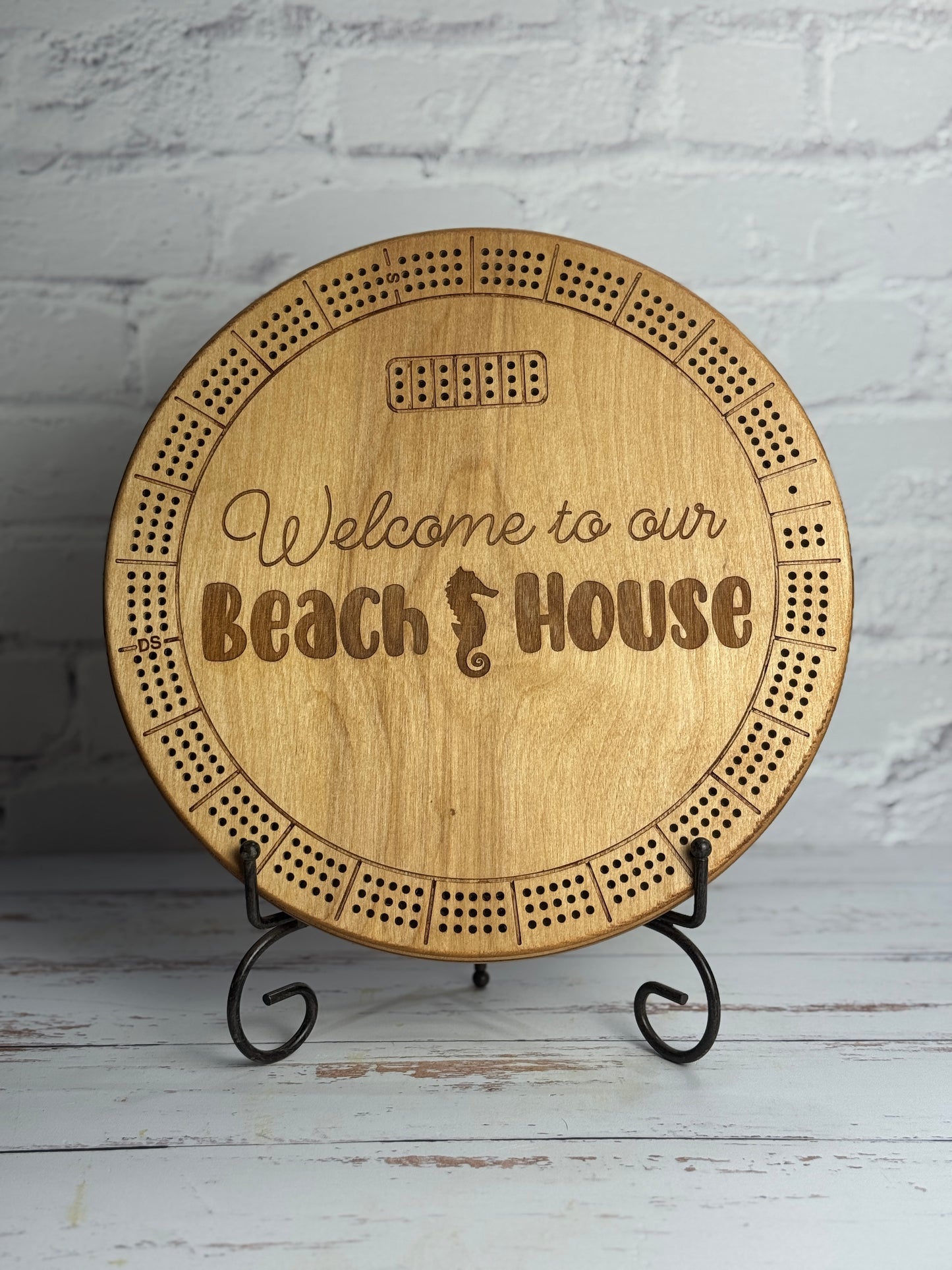 12" round light brown cribbage with 'Welcome to our Beach House' text on a stand against a white brick wall.