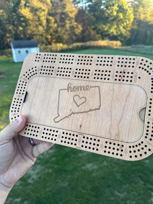 Wooden cribbage board with Connecticut 'home' state engraving held by a hand outdoors.
