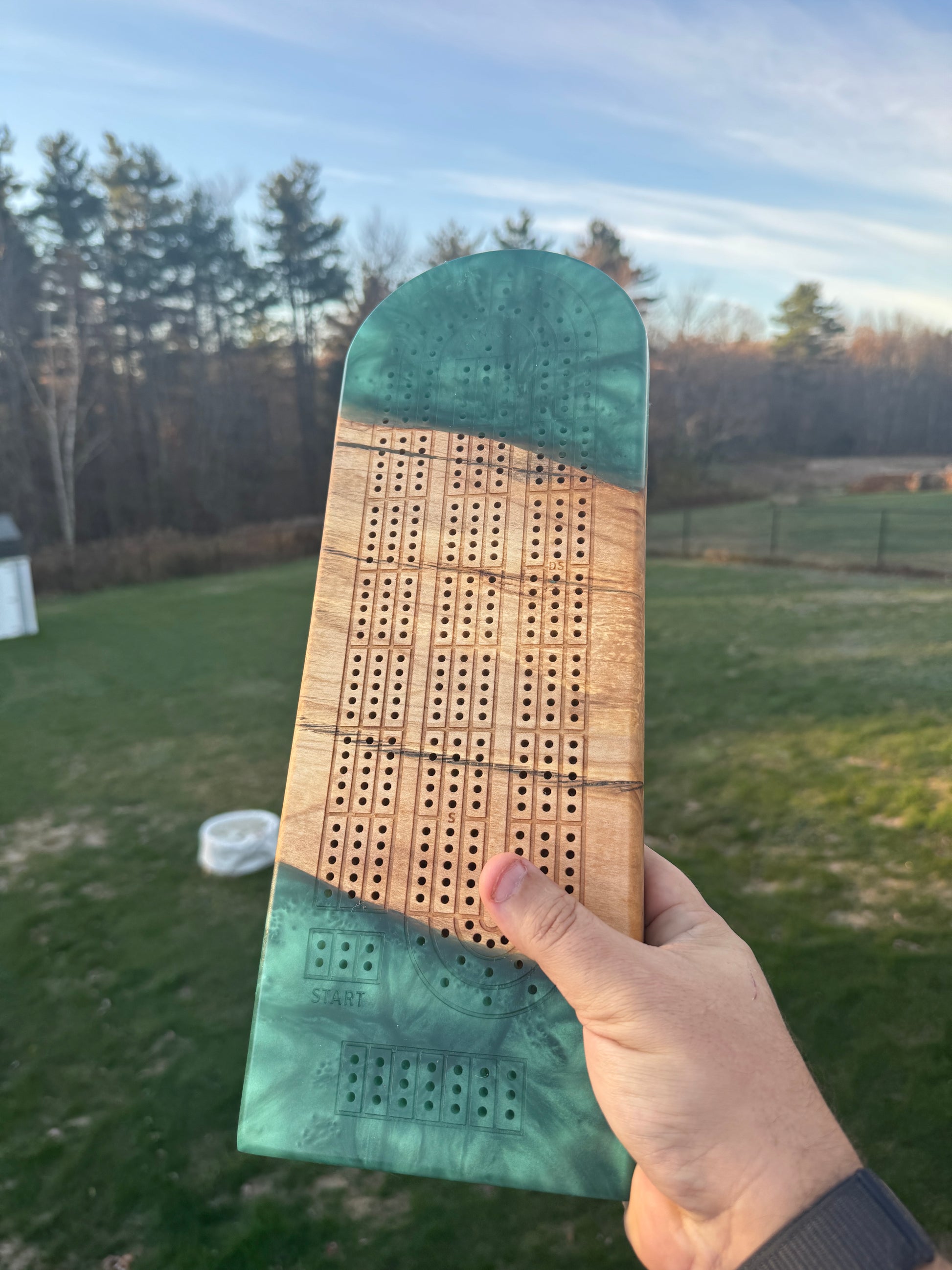 Hand holding a green resin rectangular  cribbage board, in a paperclip layout, against a nature background