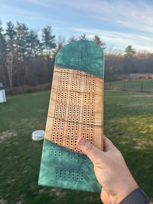 Hand holding a green resin rectangular  cribbage board, in a paperclip layout, against a nature background
